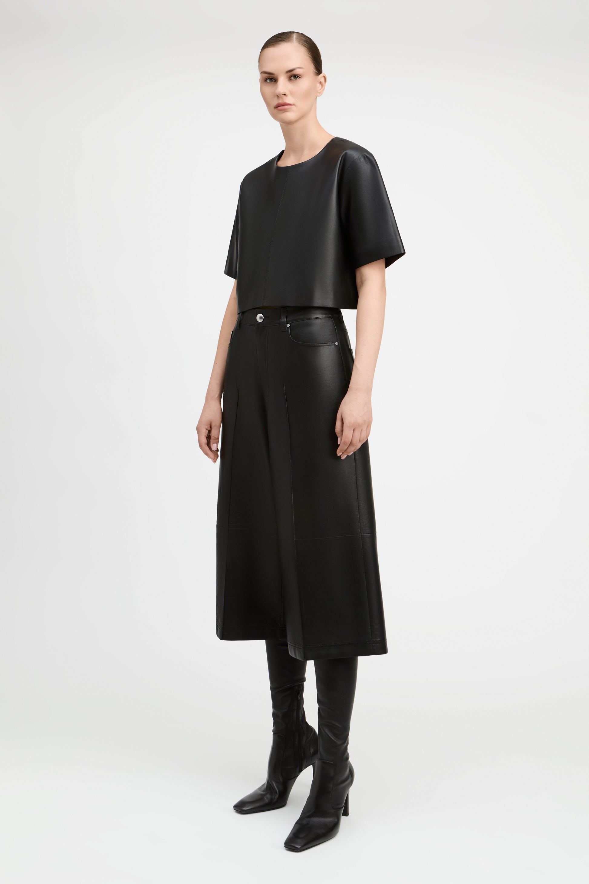 A model stands against a light gray studio background wearing a black short-sleeve cropped top, black high-waisted wide-leg skirt, black skinny boots, and black leather-look belt with a silver buckle.