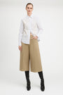 A model stands against a light gray studio background wearing a white long-sleeve button-up shirt with a heavily textured, raised floral or 3D embellishment across the chest, paired with wide-leg cropped tan pants and black knee-high boots.