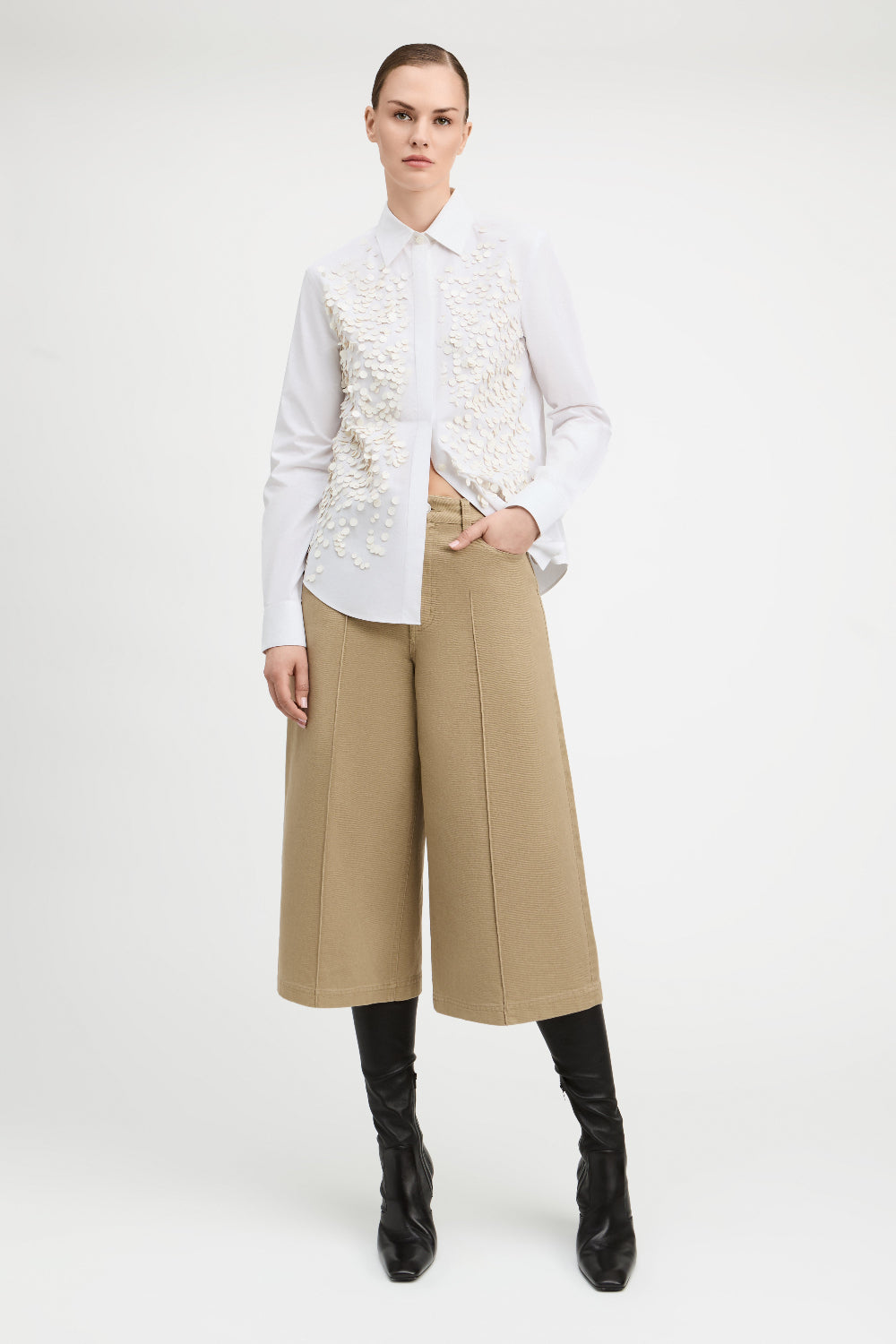 A model stands against a light gray studio background wearing a white long-sleeve button-up shirt with a heavily textured, raised floral or 3D embellishment across the chest, paired with wide-leg cropped tan pants and black knee-high boots.
