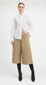 A model stands against a light gray studio background wearing a white long-sleeve button-up shirt with a heavily textured, raised floral or 3D embellishment across the chest, paired with wide-leg cropped tan pants and black knee-high boots.