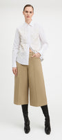 A model stands against a light gray studio background wearing a white long-sleeve button-up shirt with a heavily textured, raised floral or 3D embellishment across the chest, paired with wide-leg cropped tan pants and black knee-high boots.