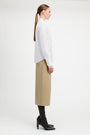 A woman in side profile wearing a white long-sleeve blouse with intricate lace embroidery on the front, paired with a beige midi skirt and black heeled boots, against a plain white studio background.