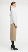 A woman in side profile wearing a white long-sleeve blouse with intricate lace embroidery on the front, paired with a beige midi skirt and black heeled boots, against a plain white studio background.