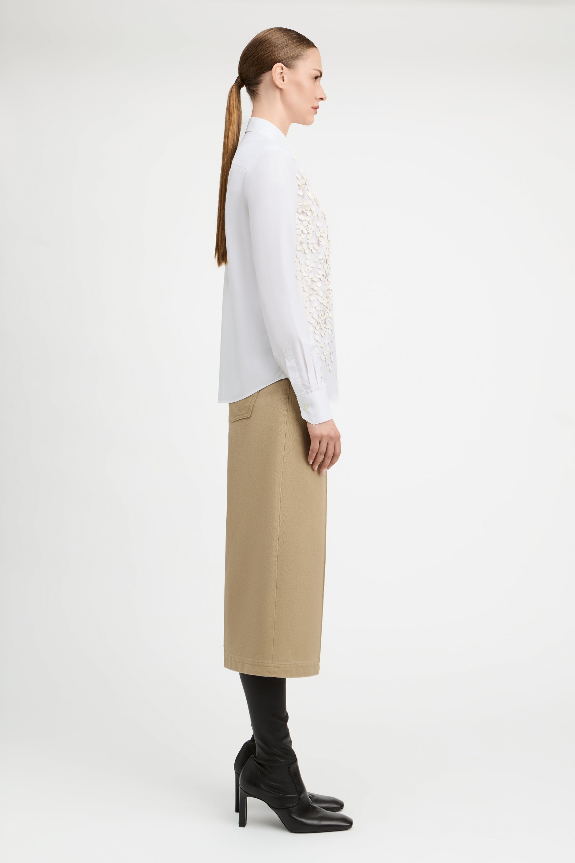 A woman in side profile wearing a white long-sleeve blouse with intricate lace embroidery on the front, paired with a beige midi skirt and black heeled boots, against a plain white studio background.