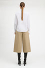 A model shown from the back wearing a white long-sleeve blouse, beige wide-leg cropped trousers, and black heeled boots against a plain white studio background.