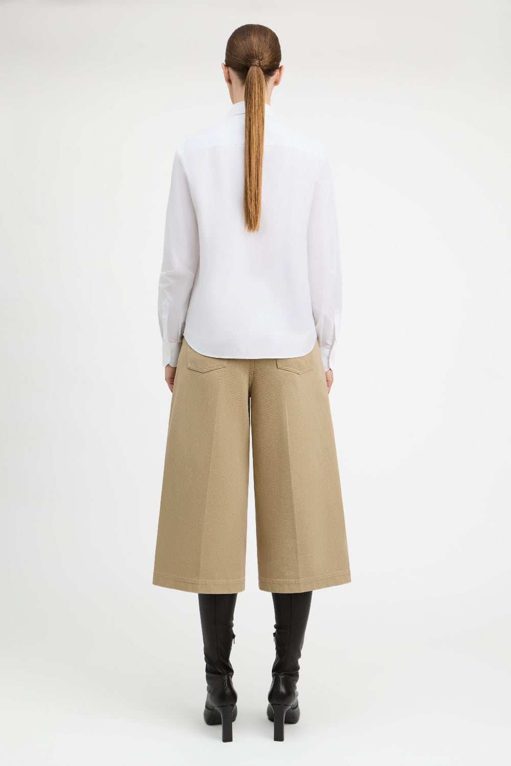A model shown from the back wearing a white long-sleeve blouse, beige wide-leg cropped trousers, and black heeled boots against a plain white studio background.