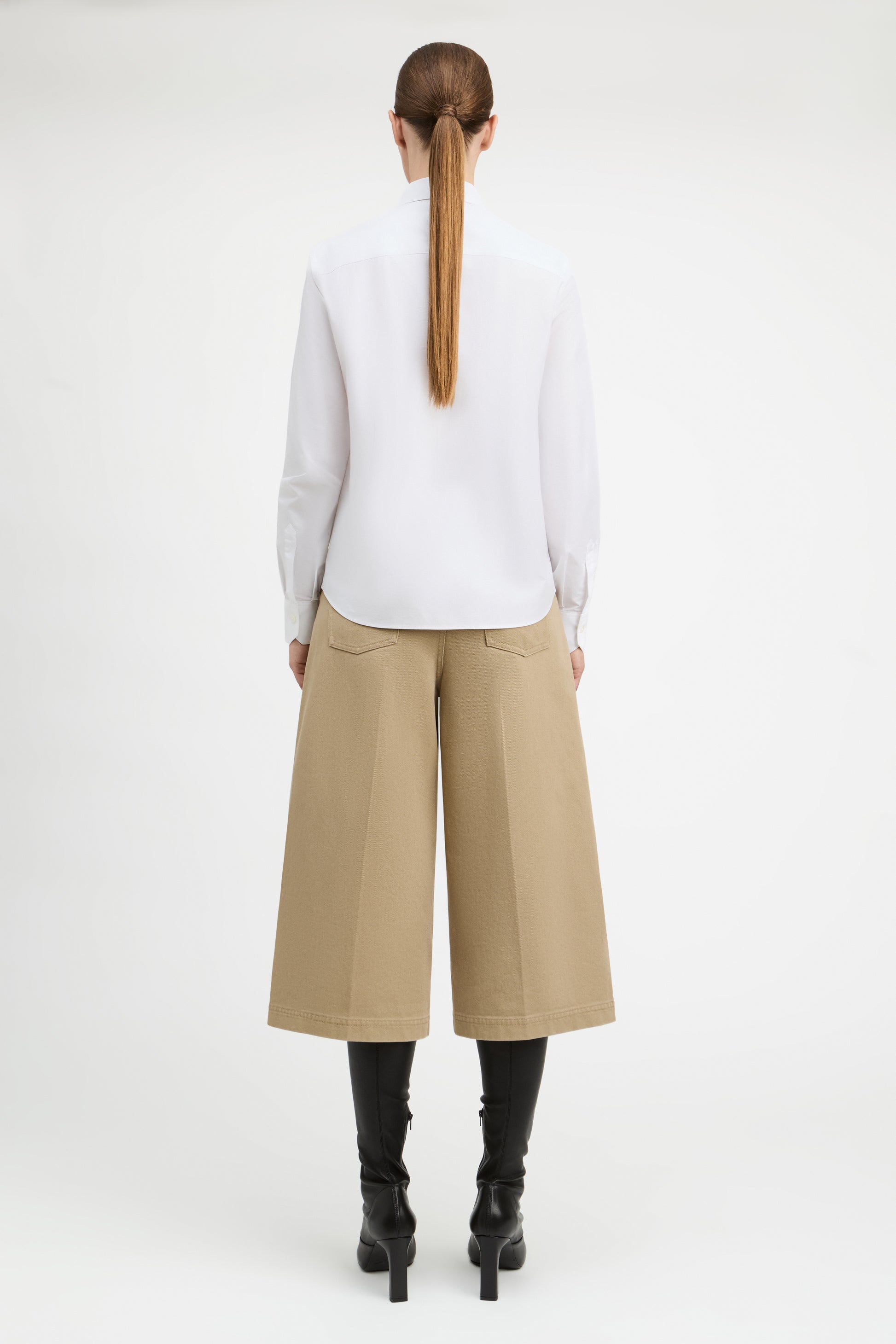 A model shown from the back wearing a white long-sleeve blouse, beige wide-leg cropped trousers, and black heeled boots against a plain white studio background.