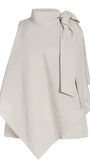 An asymmetrical cream-colored cape top with a high mock neck, textured diagonal crinkle pattern, and a large side bow detail, shown on a plain white background.