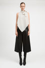 A model stands against a light gray studio background wearing a sleeveless high-neck wrap top in off-white with a bow detail at the collar, paired with wide-leg black pants and black knee-high boots.