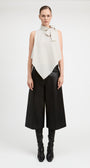 A model stands against a light gray studio background wearing a sleeveless high-neck wrap top in off-white with a bow detail at the collar, paired with wide-leg black pants and black knee-high boots.