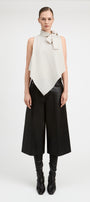 A model stands against a light gray studio background wearing a sleeveless high-neck wrap top in off-white with a bow detail at the collar, paired with wide-leg black pants and black knee-high boots.