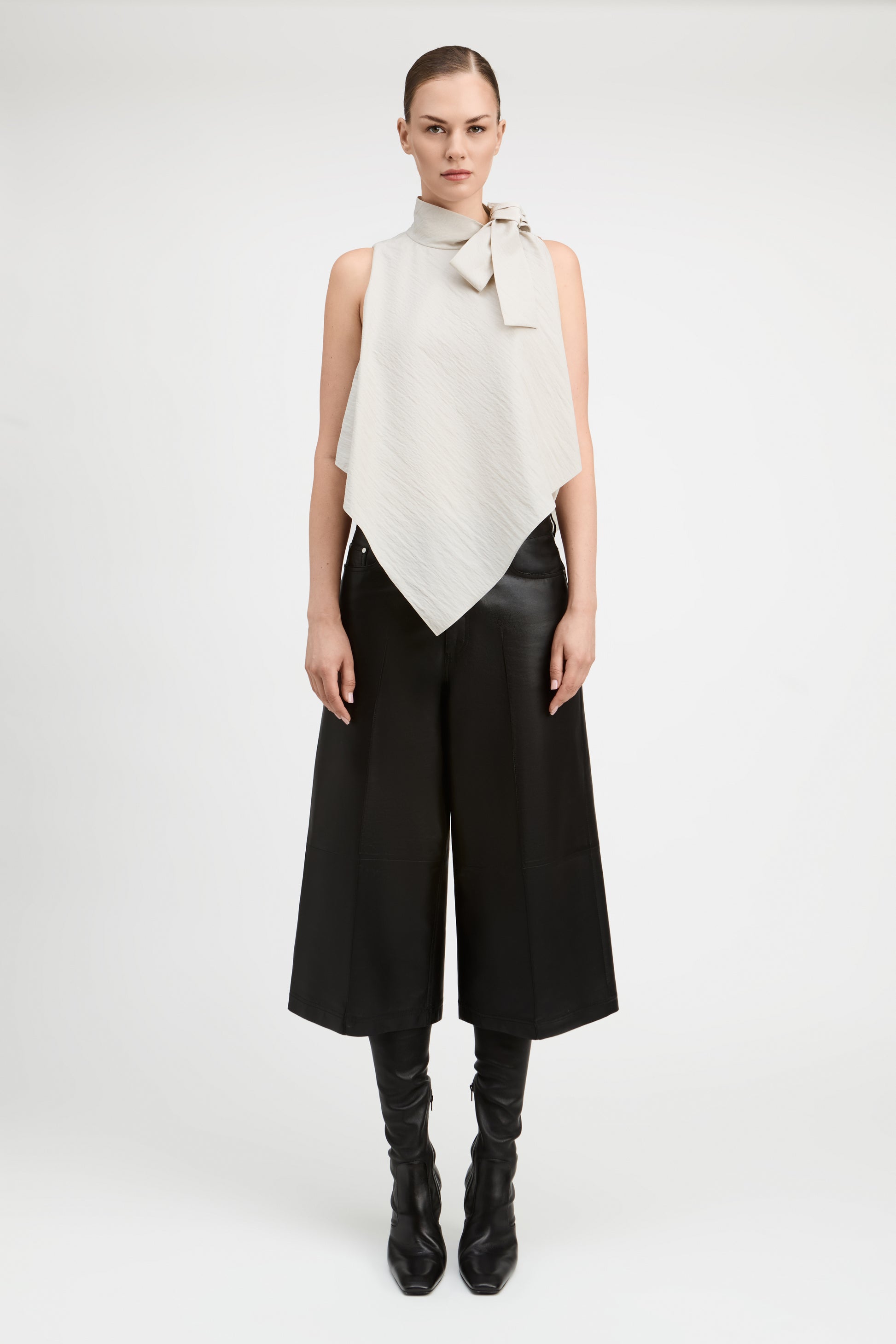 A model stands against a light gray studio background wearing a sleeveless high-neck wrap top in off-white with a bow detail at the collar, paired with wide-leg black pants and black knee-high boots.