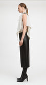 A model is shown in profile wearing a sleeveless cream draped top, a black midi skirt, and black heeled boots against a plain white studio background.