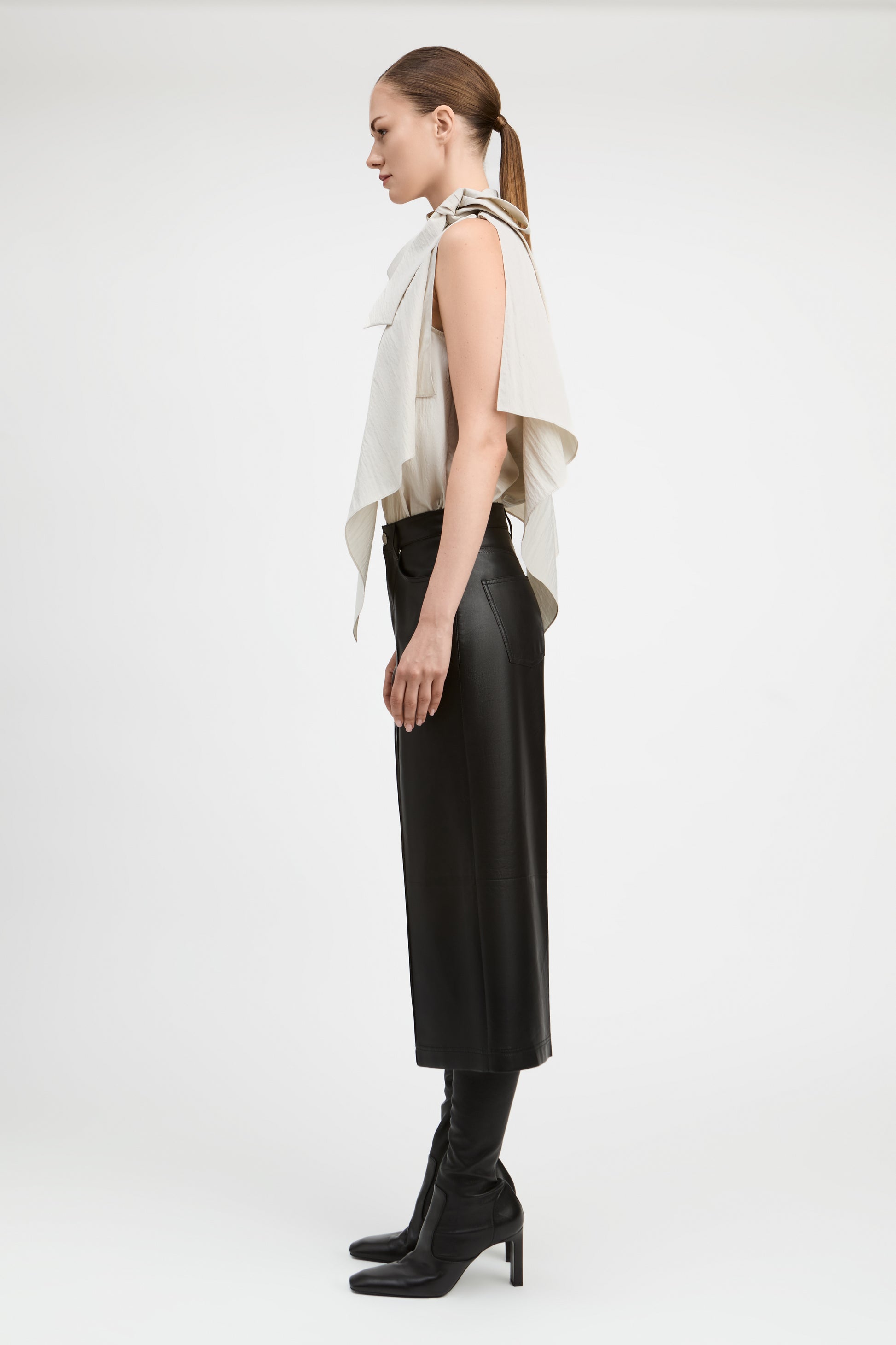 A model is shown in profile wearing a sleeveless cream draped top, a black midi skirt, and black heeled boots against a plain white studio background.