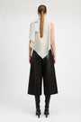 A model shown from the back wearing a sleeveless asymmetrical cream draped top, black wide-leg cropped pants, and black heeled boots against a plain light gray studio background.
