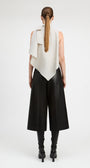 A model shown from the back wearing a sleeveless asymmetrical cream draped top, black wide-leg cropped pants, and black heeled boots against a plain light gray studio background.