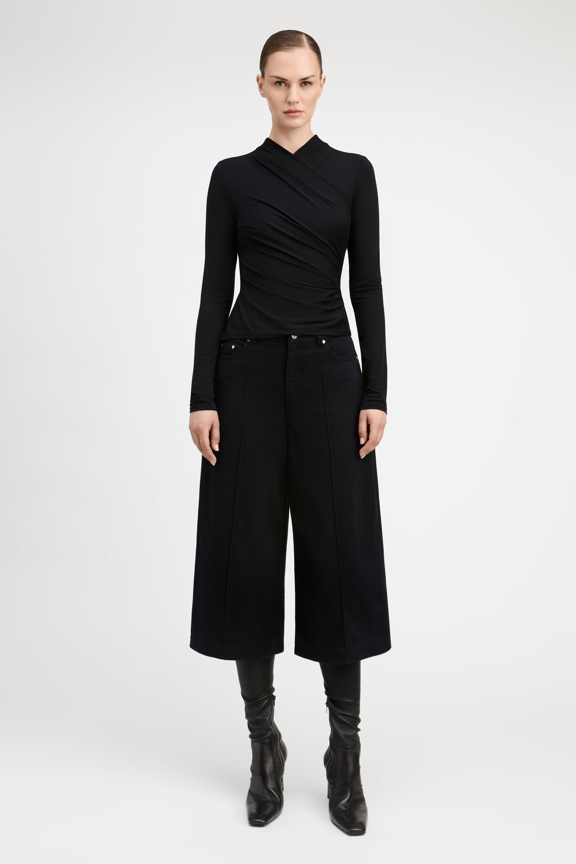 A model stands against a light gray studio background wearing a black asymmetrical long-sleeve top, black wide-leg cropped trousers, and black knee-high boots.