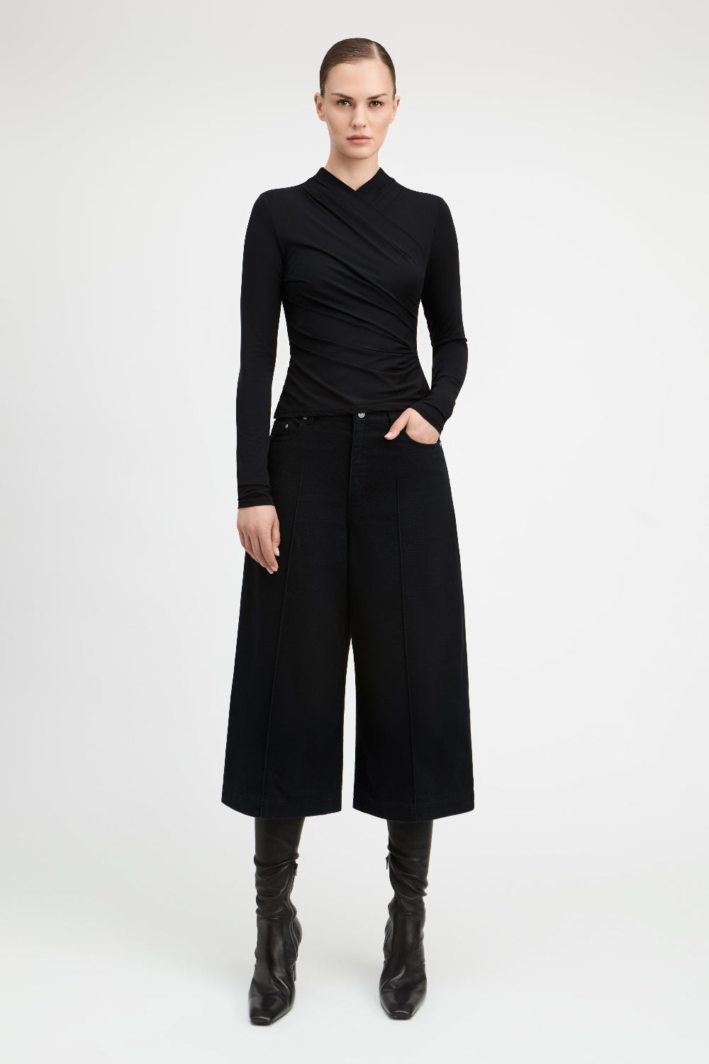 A model stands against a light gray studio background wearing a black long-sleeve wrap-style top, black wide-leg cropped pants, and black knee-high boots.