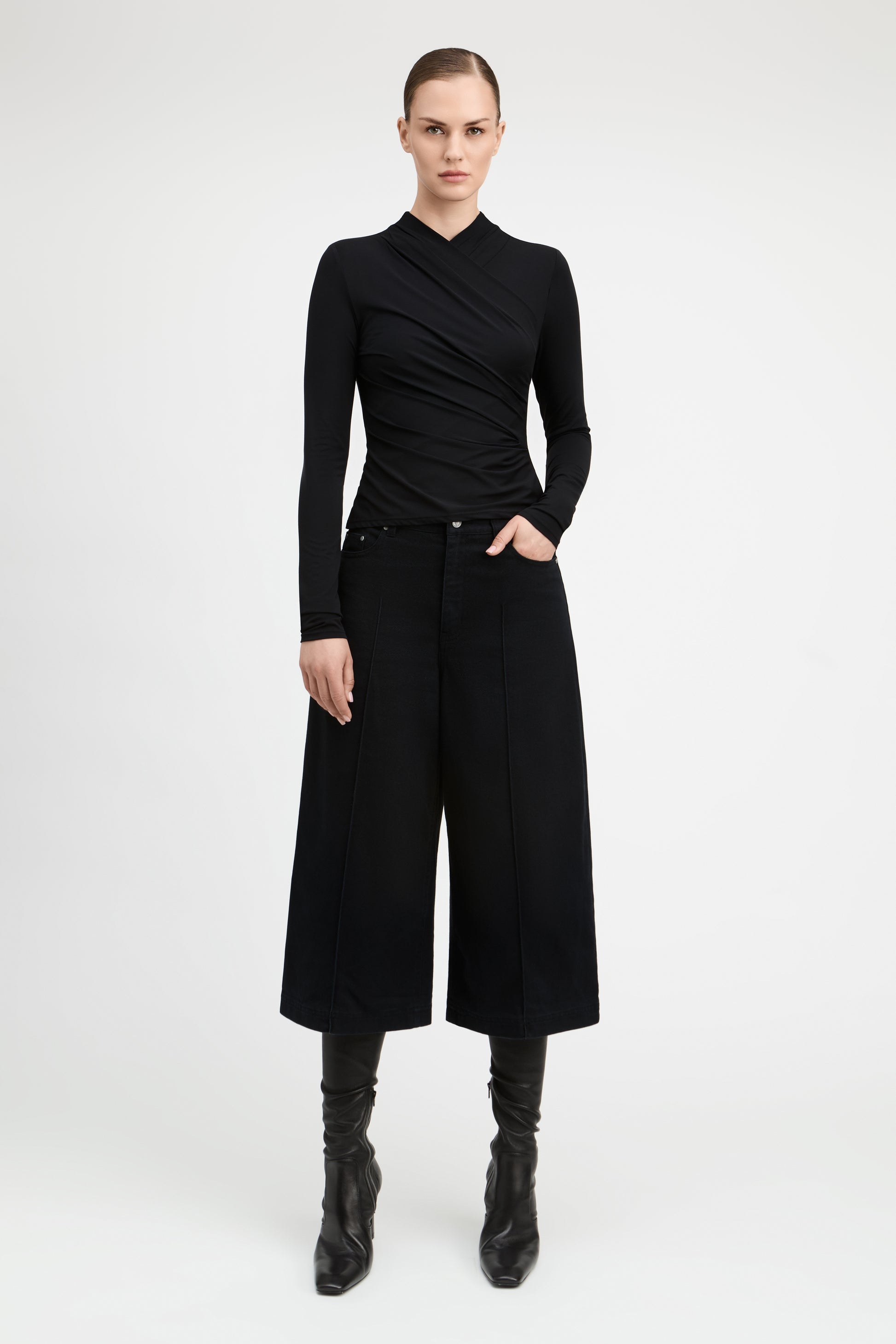 A model stands against a light gray studio background wearing a black long-sleeve wrap-style top, black wide-leg cropped pants, and black knee-high boots.