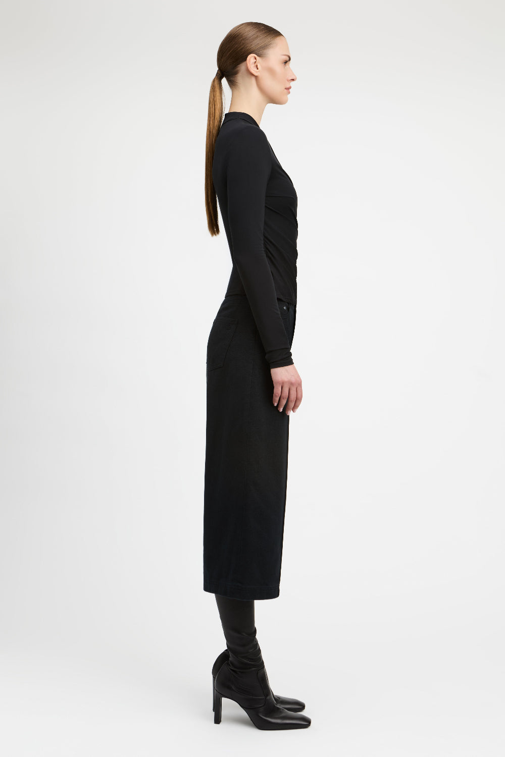 A woman in a side profile wearing a long-sleeve black midi dress, black knee-high socks, and black high-heeled boots against a plain light gray studio background.