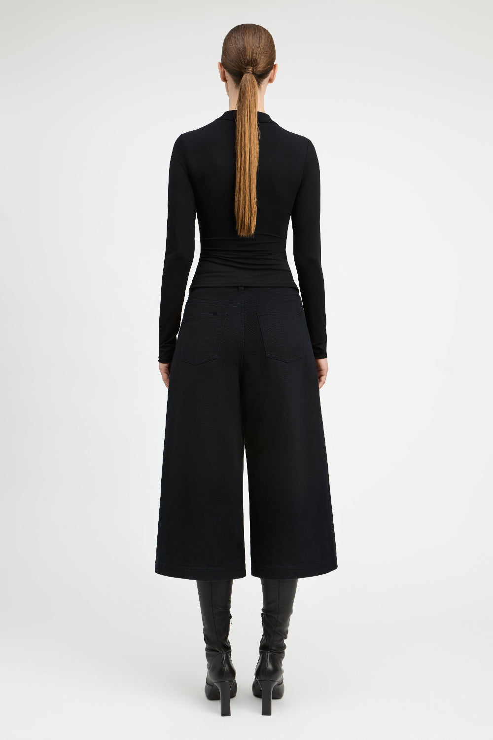 A model viewed from the back wearing a black long-sleeve top and wide-leg cropped pants, paired with black heeled boots, against a plain light gray studio background.