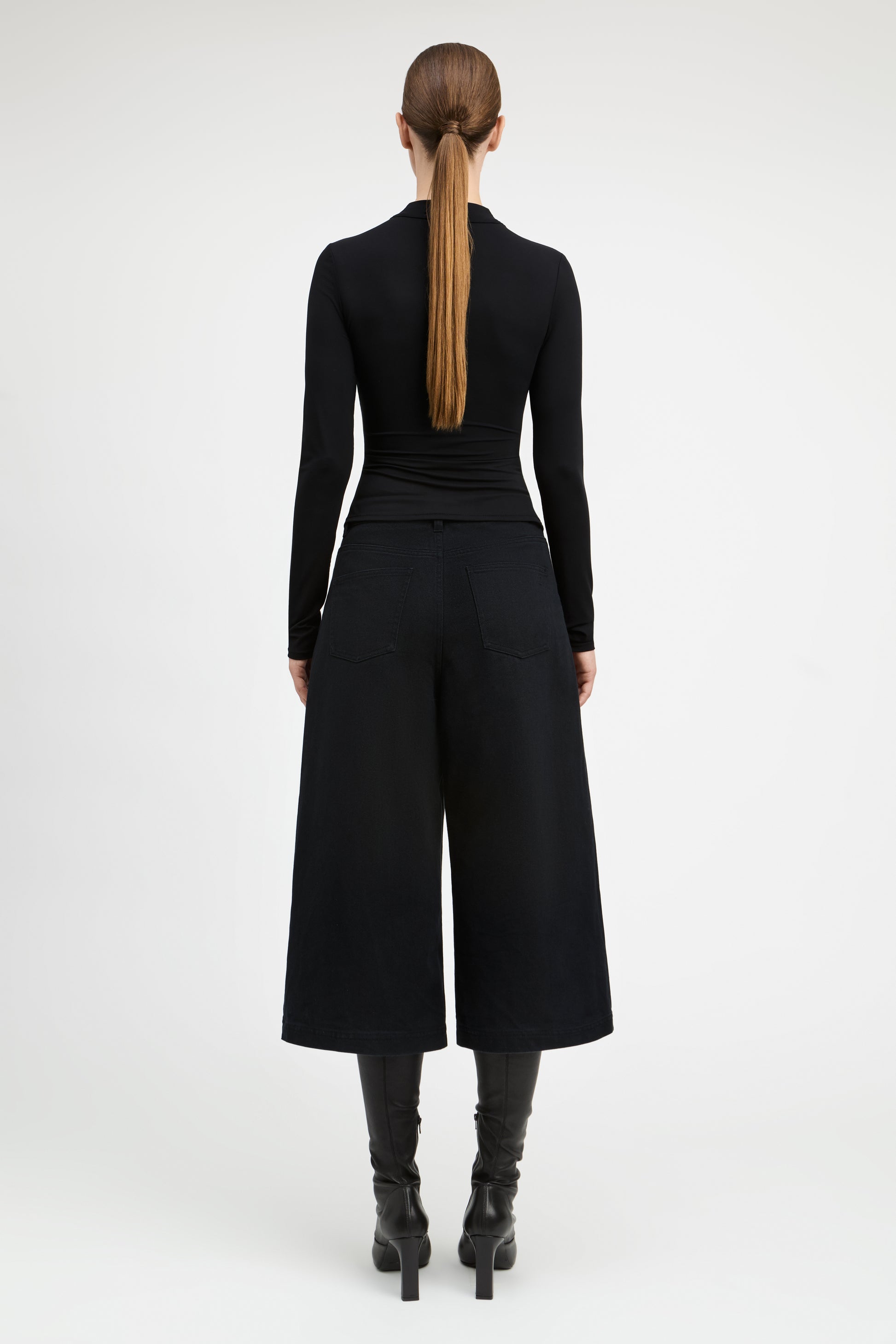 A model viewed from the back wearing a black long-sleeve top and wide-leg cropped pants, paired with black heeled boots, against a plain light gray studio background.