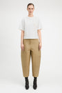 A model stands against a light gray studio background wearing a white short-sleeve top with textured embroidery, beige wide-leg cropped pants with side pockets, and black ankle boots.