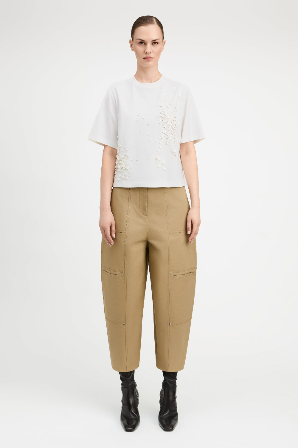 A model stands against a light gray studio background wearing a white short-sleeve top with textured embroidery, beige wide-leg cropped pants with side pockets, and black ankle boots.