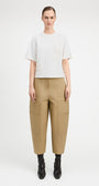 A model stands against a light gray studio background wearing a white short-sleeve top with textured embroidery, beige wide-leg cropped pants with side pockets, and black ankle boots.