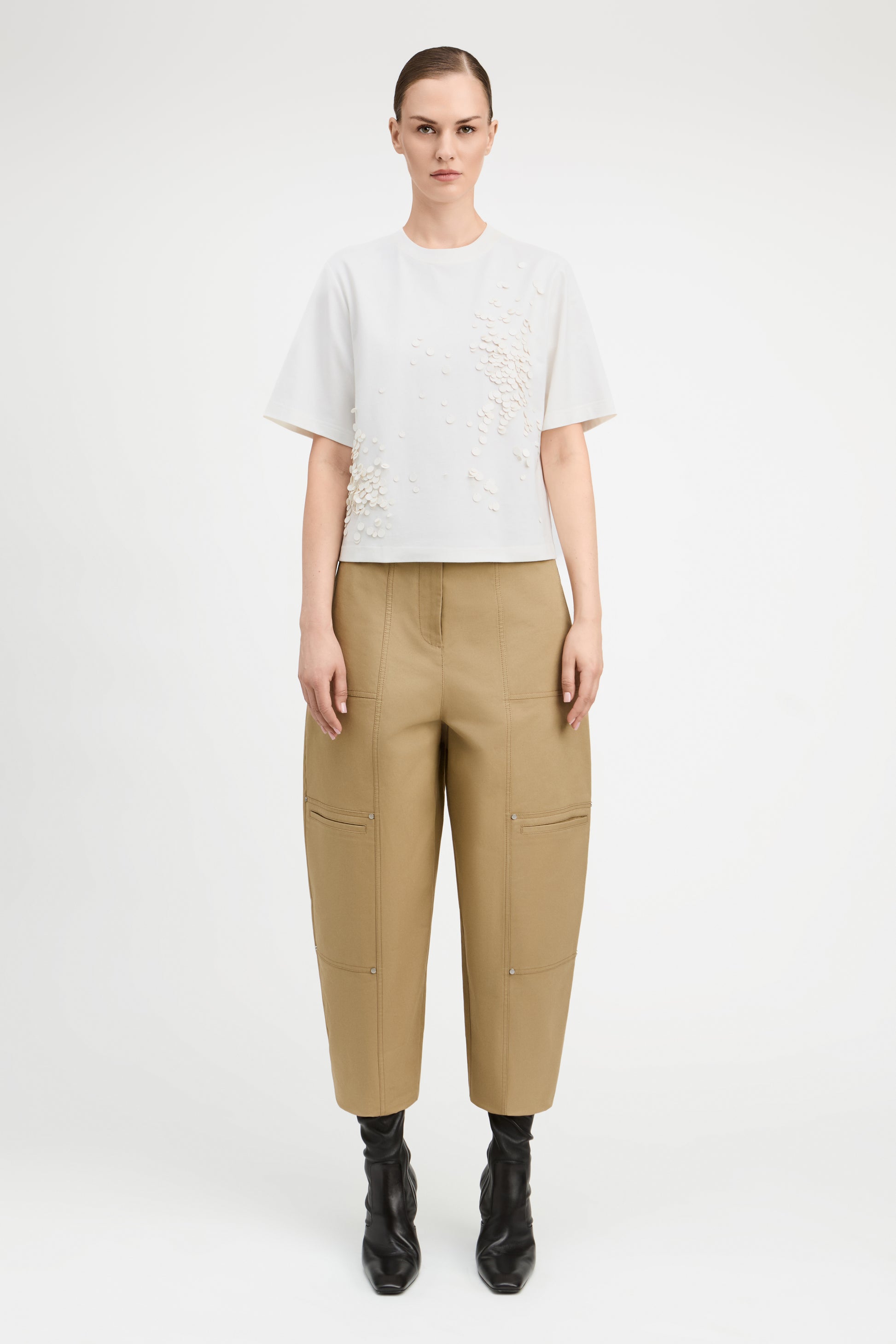 A model stands against a light gray studio background wearing a white short-sleeve top with textured embroidery, beige wide-leg cropped pants with side pockets, and black ankle boots.