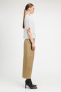 A woman in profile wearing a white short-sleeve top with textured side panels and beige wide-leg cropped pants, paired with black ankle boots, against a plain light gray studio background.