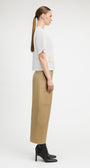 A woman in profile wearing a white short-sleeve top with textured side panels and beige wide-leg cropped pants, paired with black ankle boots, against a plain light gray studio background.