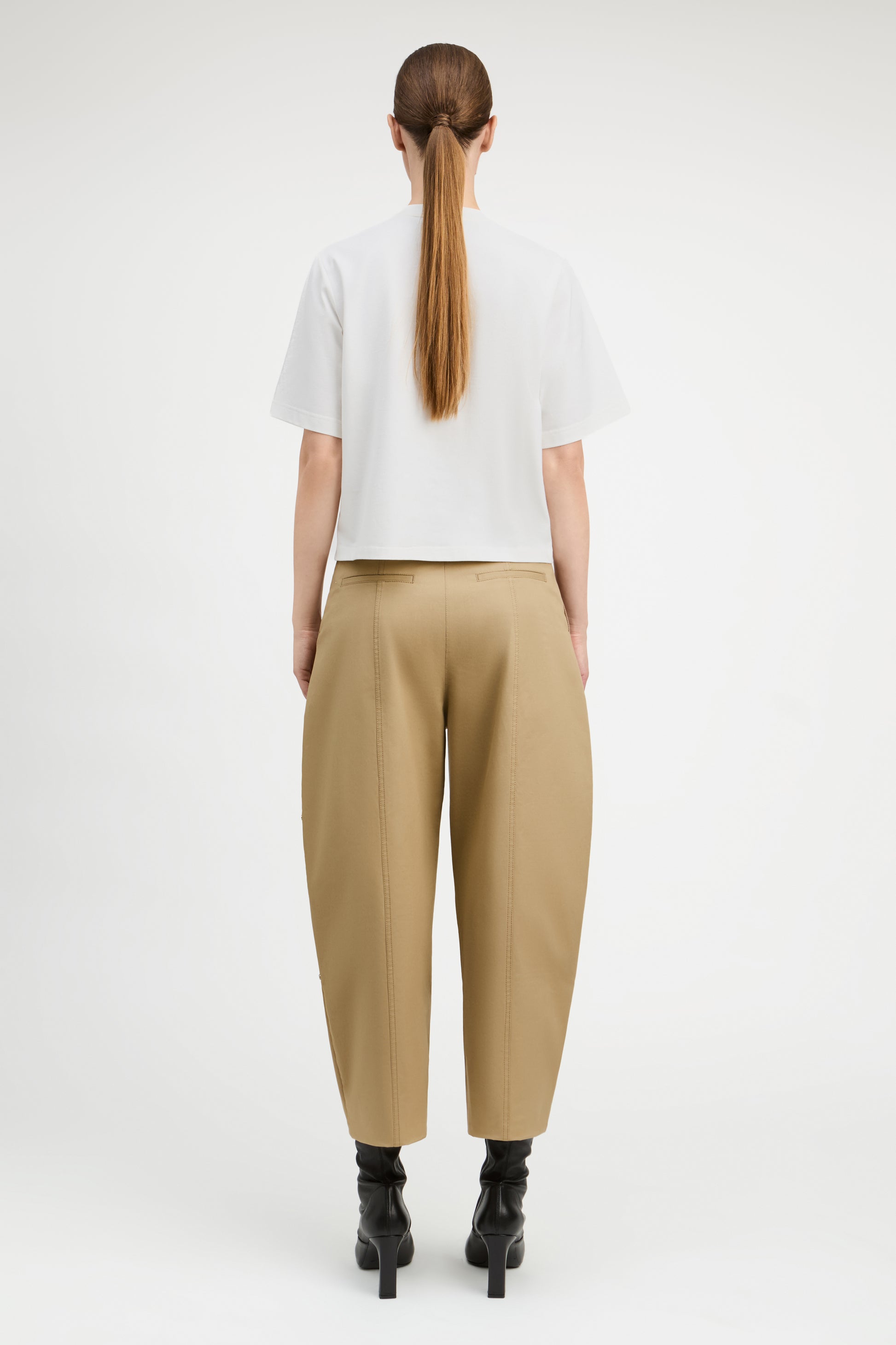 A model viewed from the back wearing a white short-sleeve cropped T-shirt, beige wide-leg cropped trousers, and black ankle boots against a plain light gray studio background.