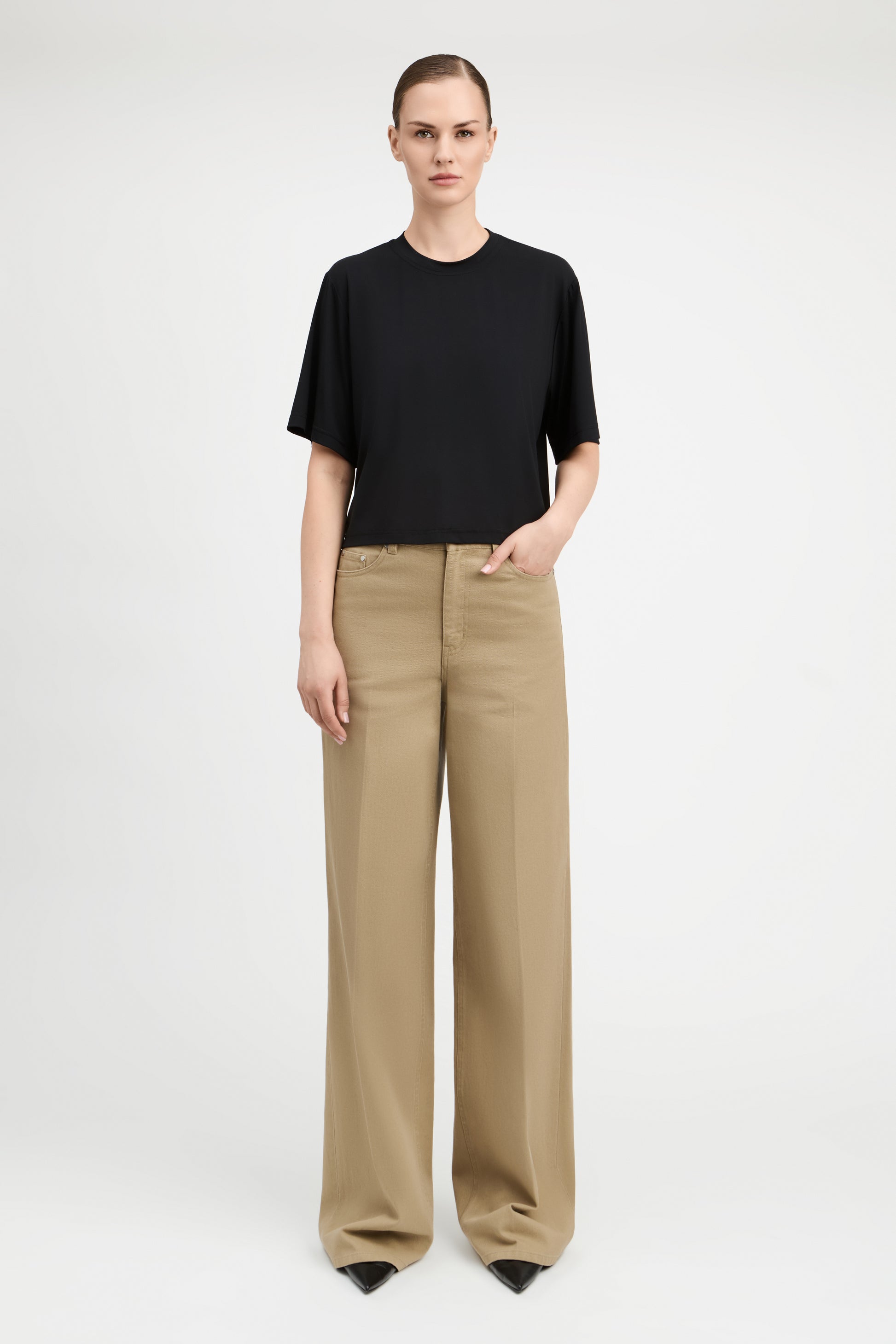 A model stands against a light gray studio background wearing a black short-sleeve crewneck top and beige wide-leg trousers.