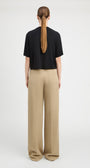 A woman viewed from the back wearing a black short-sleeve cropped T-shirt and beige wide-leg trousers, standing against a plain light gray studio background.