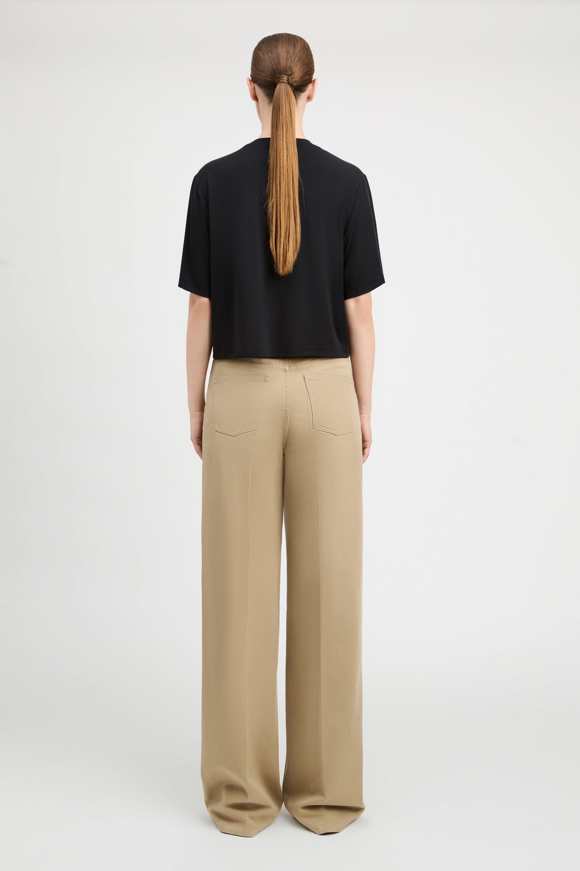 A woman viewed from the back wearing a black short-sleeve cropped T-shirt and beige wide-leg trousers, standing against a plain light gray studio background.