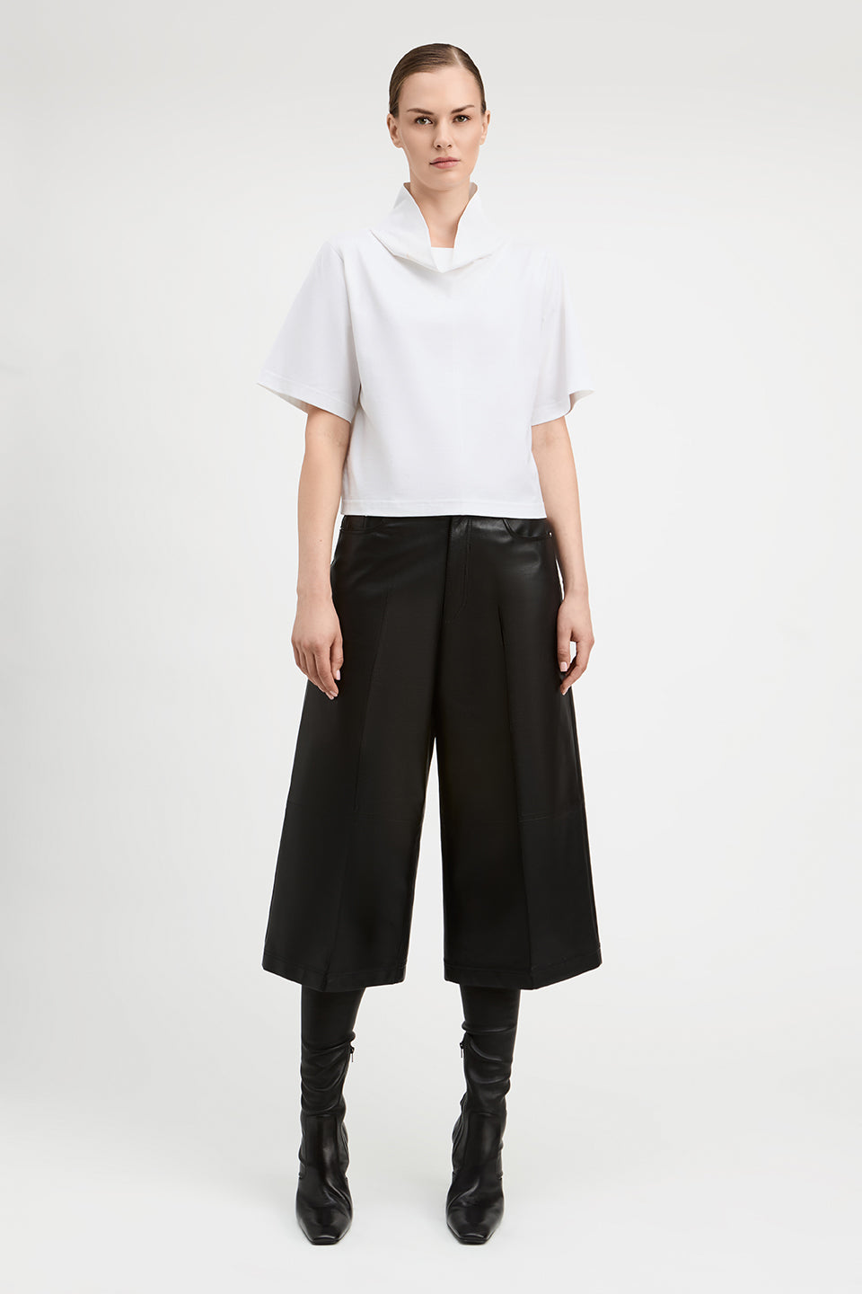 A model stands against a light gray studio background wearing a white short-sleeve top with a high draped neckline, black wide-leg cropped pants, and black knee-high boots.
