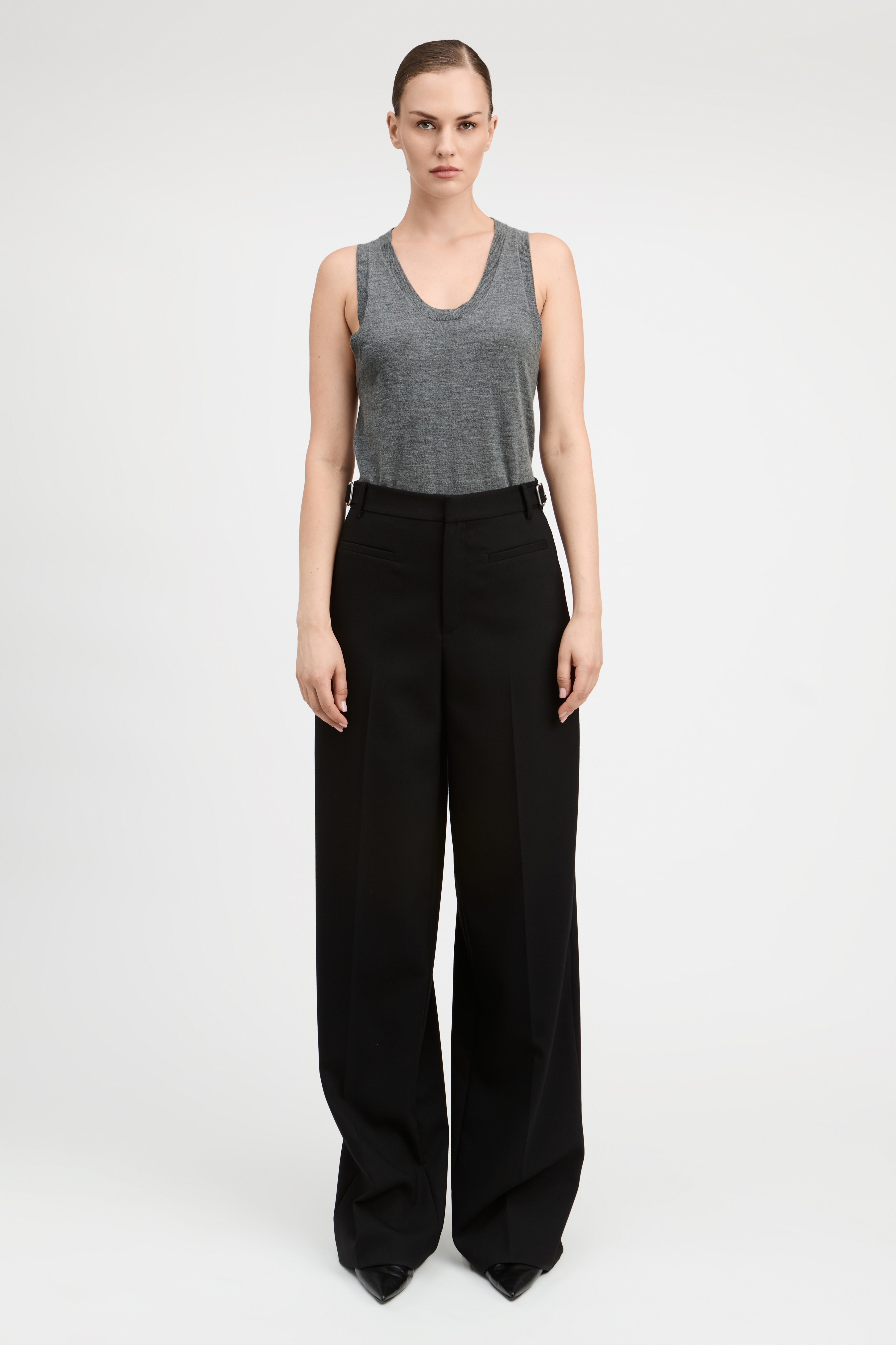 A woman wearing a gray sleeveless top and black wide-leg trousers, standing against a plain light gray studio background.