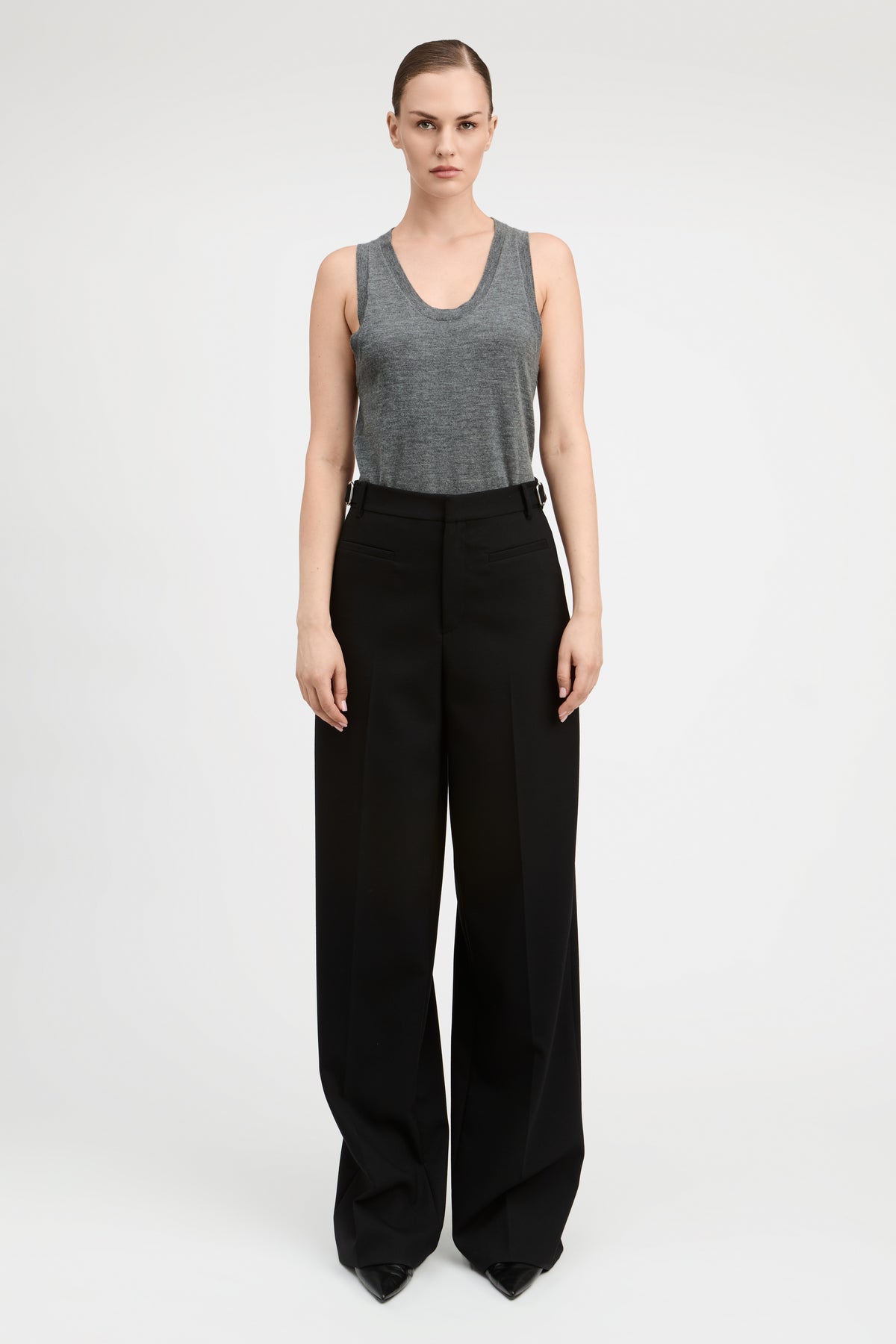 A woman wearing a gray sleeveless top and black wide-leg trousers, standing against a plain light gray studio background.