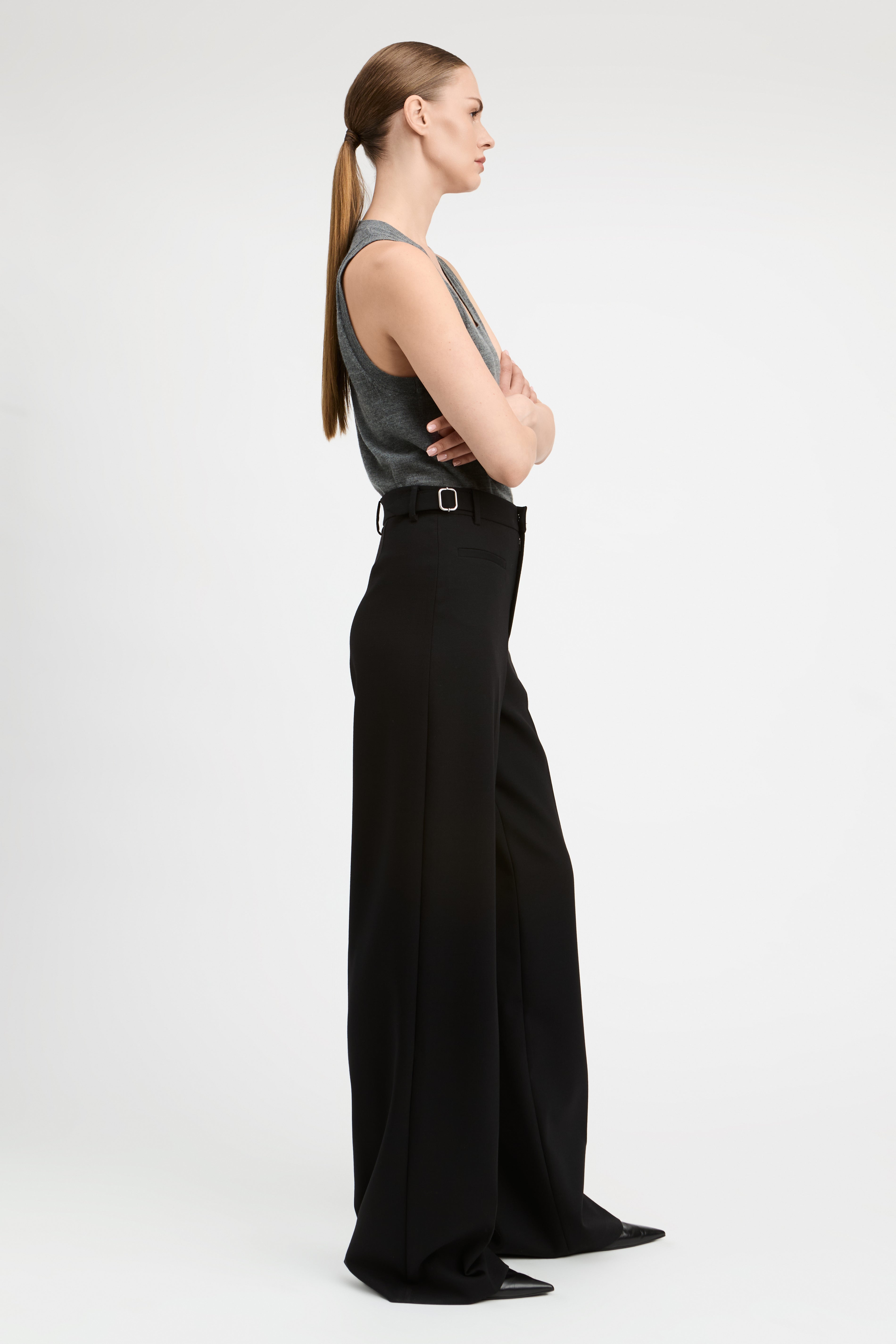 A woman on the side wearing a gray sleeveless top and black wide-leg trousers, standing against a plain light gray studio background.