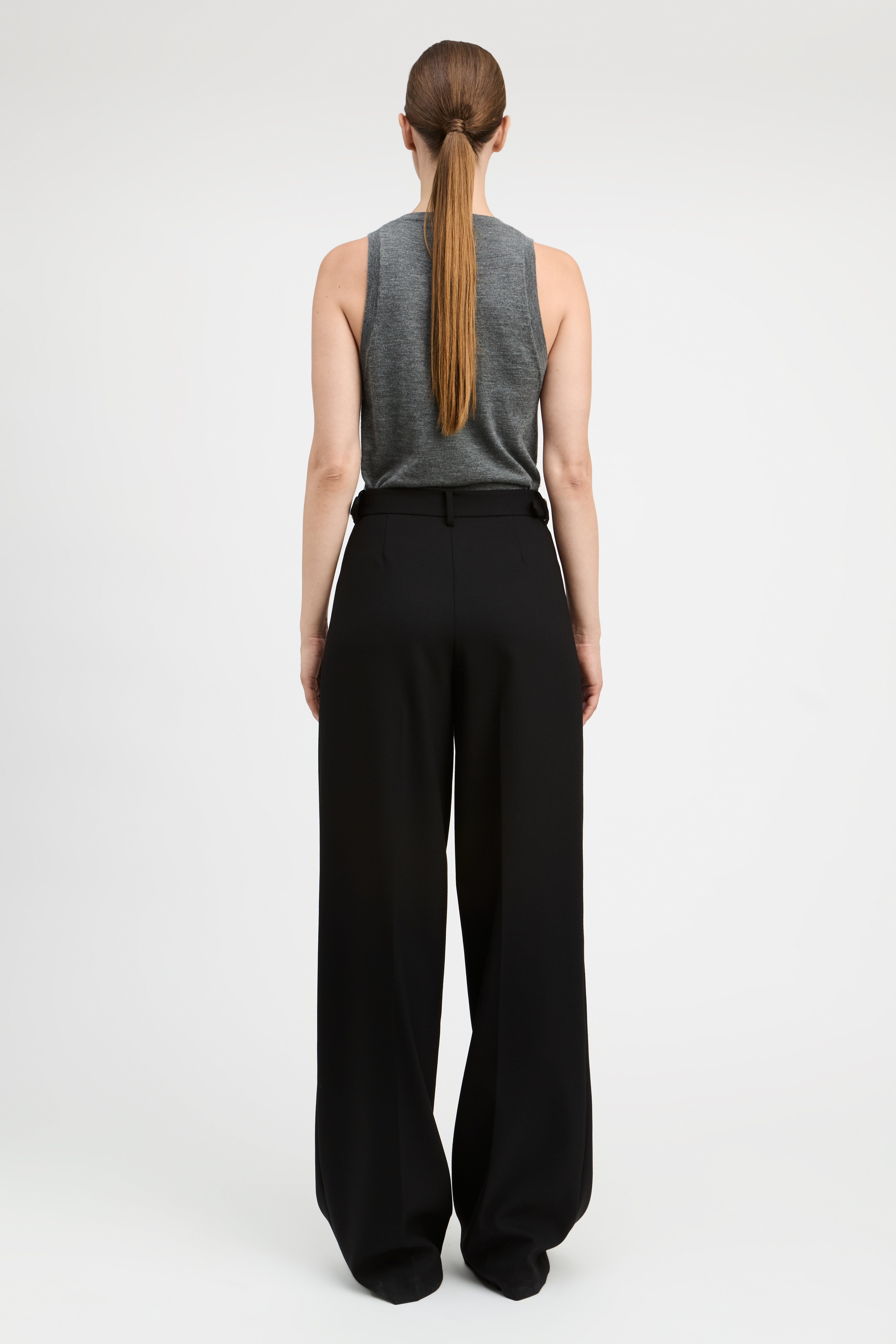 A woman viewed from the back wearing a gray sleeveless top and black wide-leg trousers, standing against a plain light gray studio background.