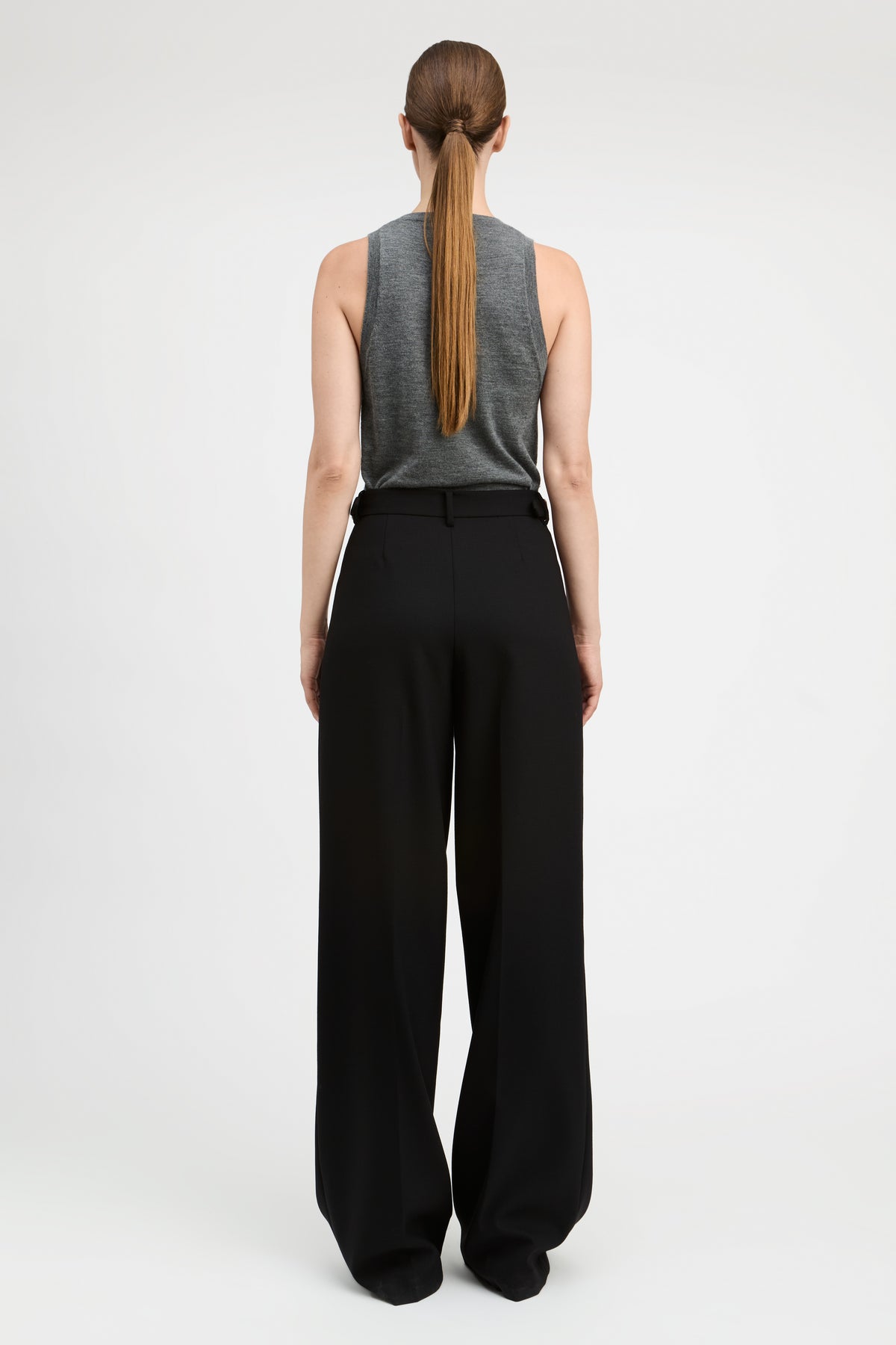 A woman viewed from the back wearing a gray sleeveless top and black wide-leg trousers, standing against a plain light gray studio background.