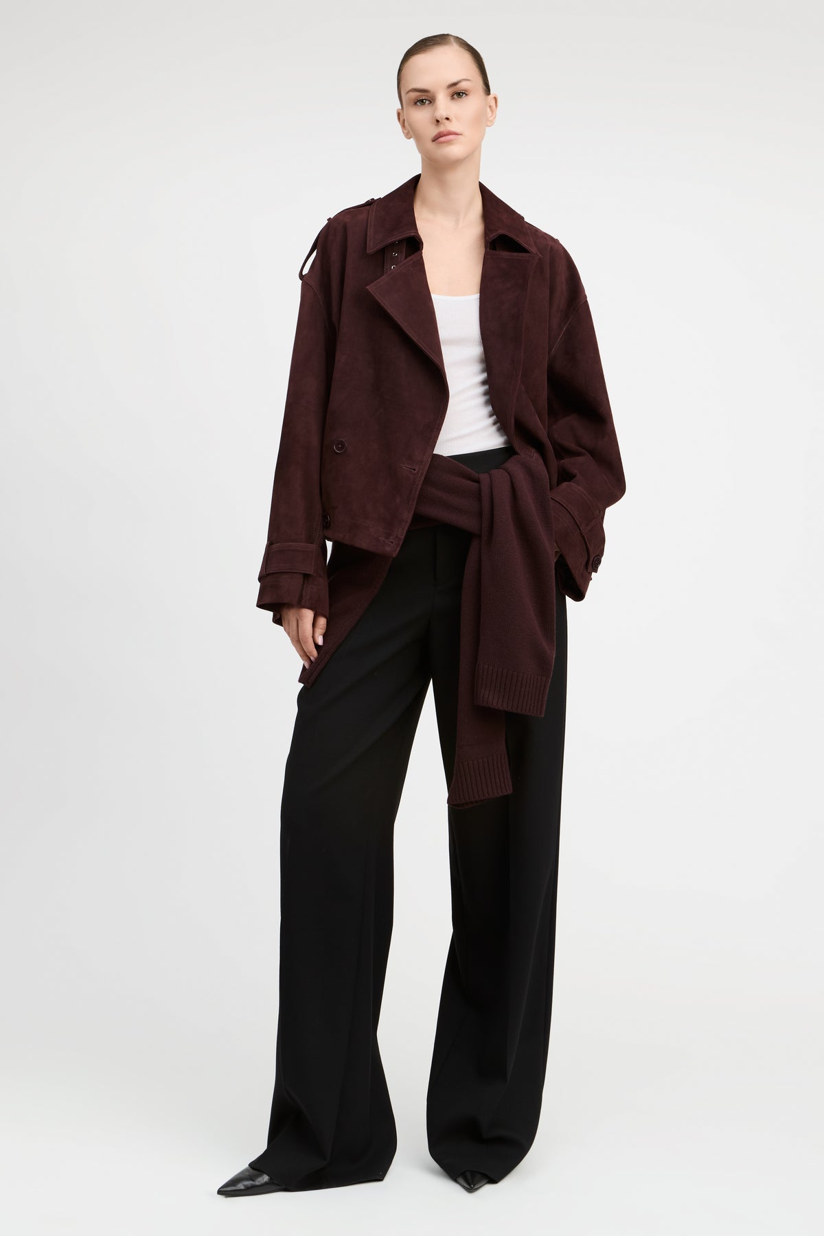 A model stands against a light gray studio background wearing a dark burgundy corduroy jacket over a white top and black wide-leg trousers.