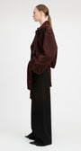 A model stands against a light gray studio background wearing a dark burgundy corduroy jacket over a white top and black wide-leg trousers.