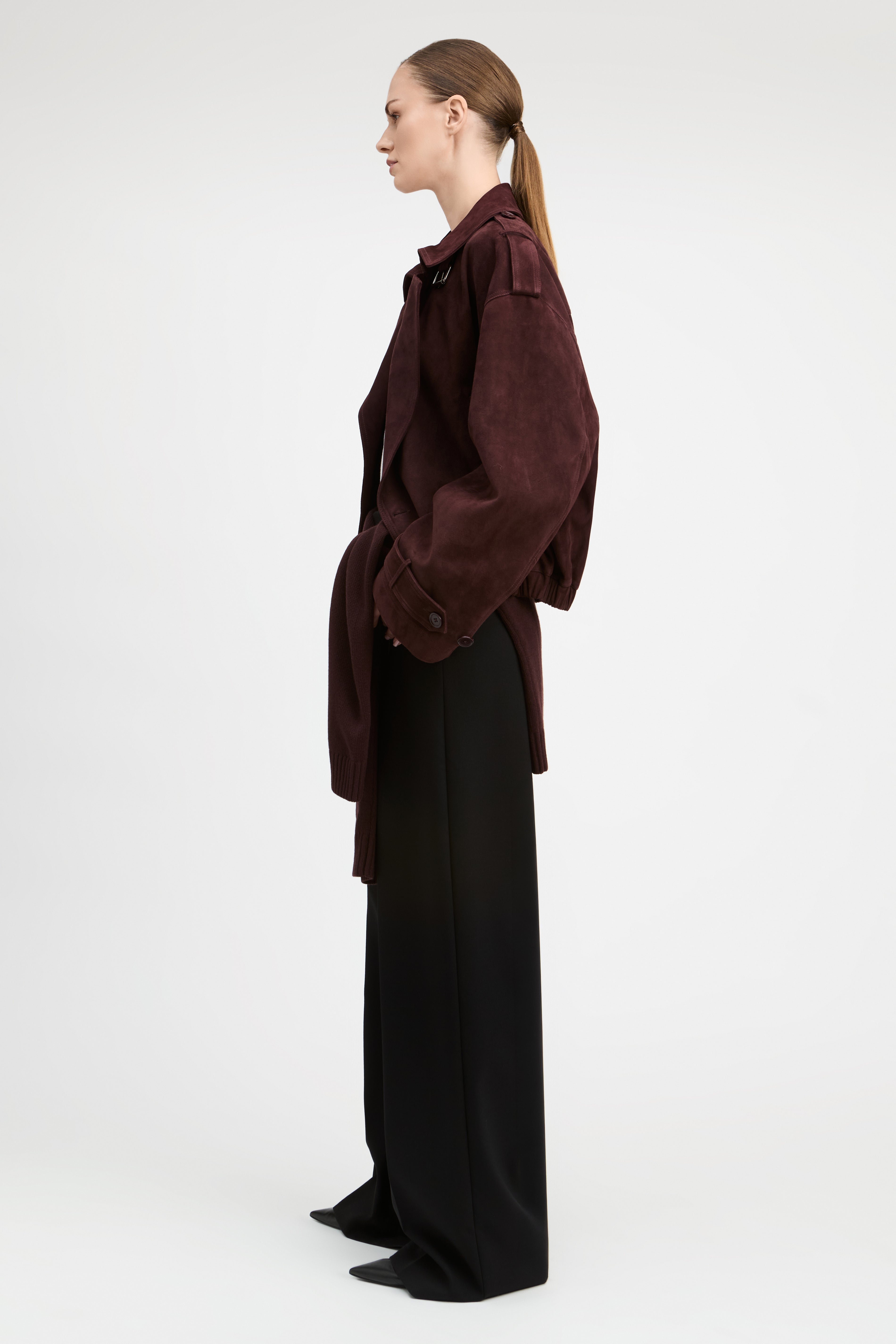 A model stands against a light gray studio background wearing a dark burgundy corduroy jacket over a white top and black wide-leg trousers.