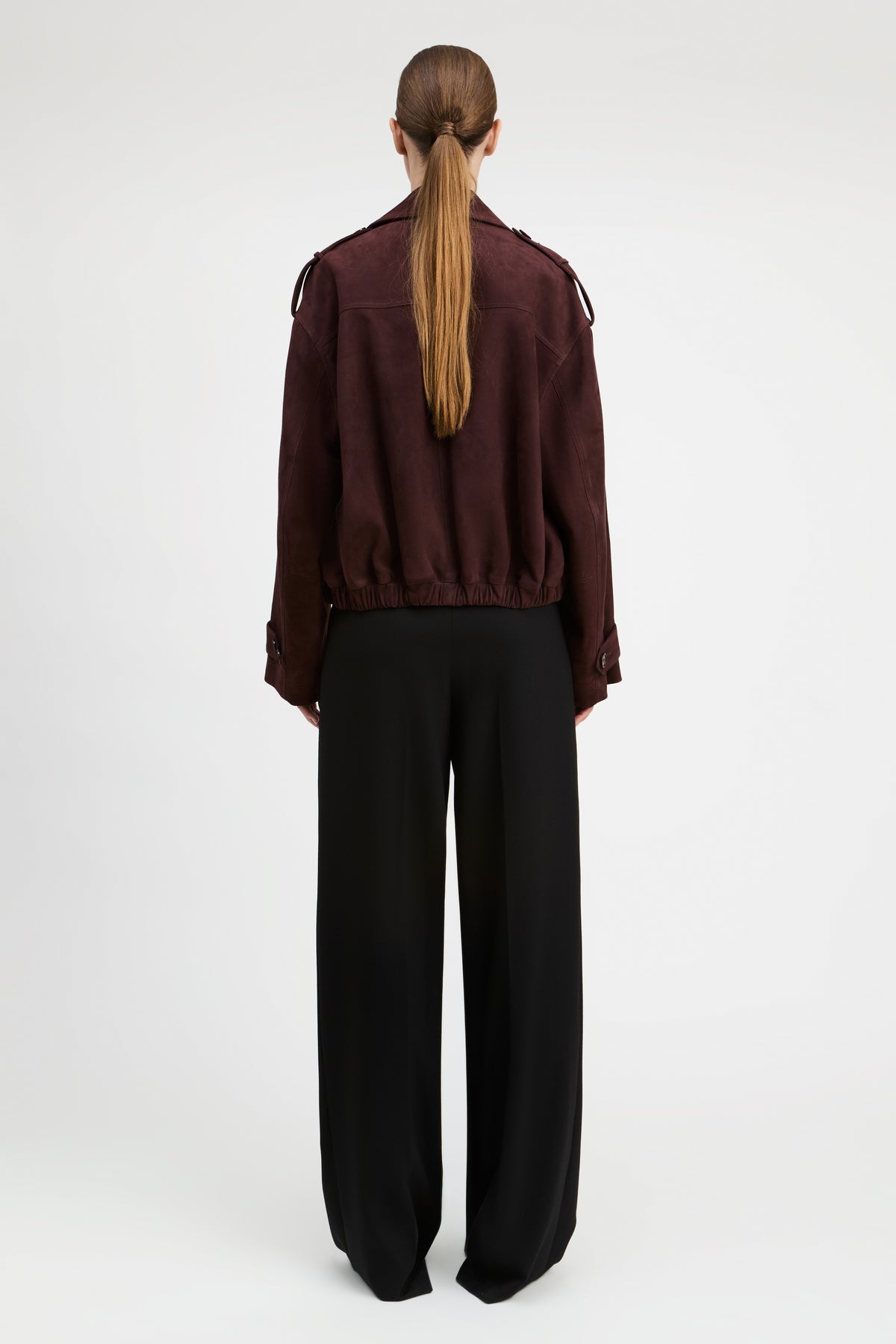 A model shown from the back wearing a dark burgundy cropped jacket with shoulder epaulets and black wide-leg trousers, set against a plain light gray studio background.