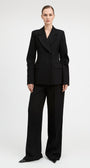 A model wearing a black double-breasted blazer and matching wide-leg trousers, photographed against a seamless light gray studio background.