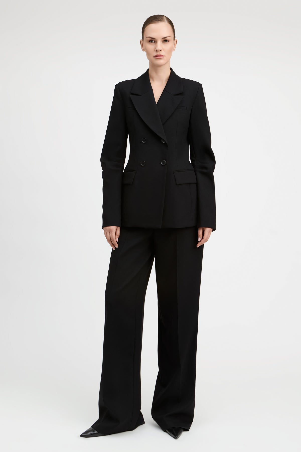 A model wearing a black double-breasted blazer and matching wide-leg trousers, photographed against a seamless light gray studio background.