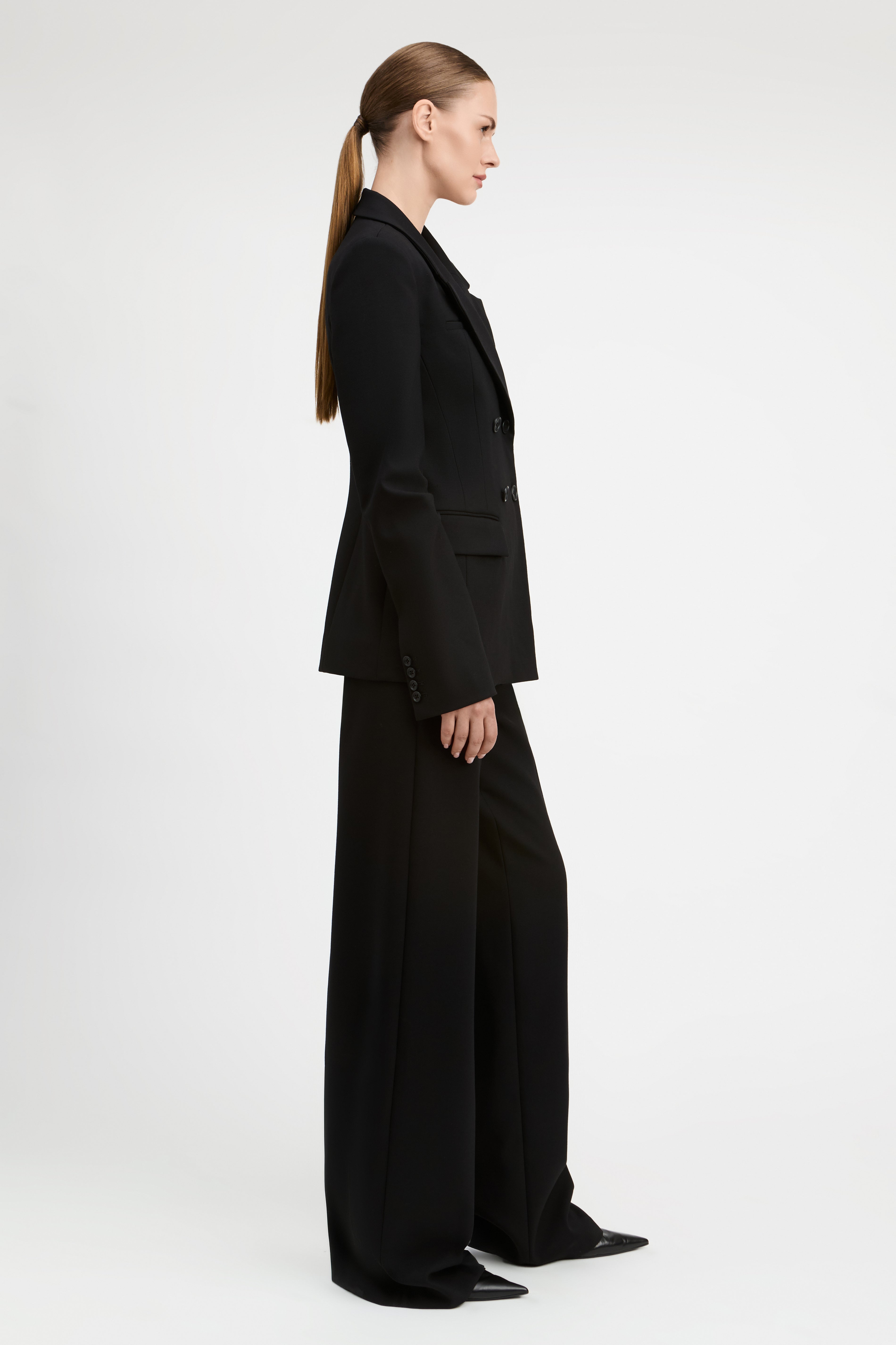 A model wearing a black double-breasted blazer and matching wide-leg trousers, photographed against a seamless light gray studio background.