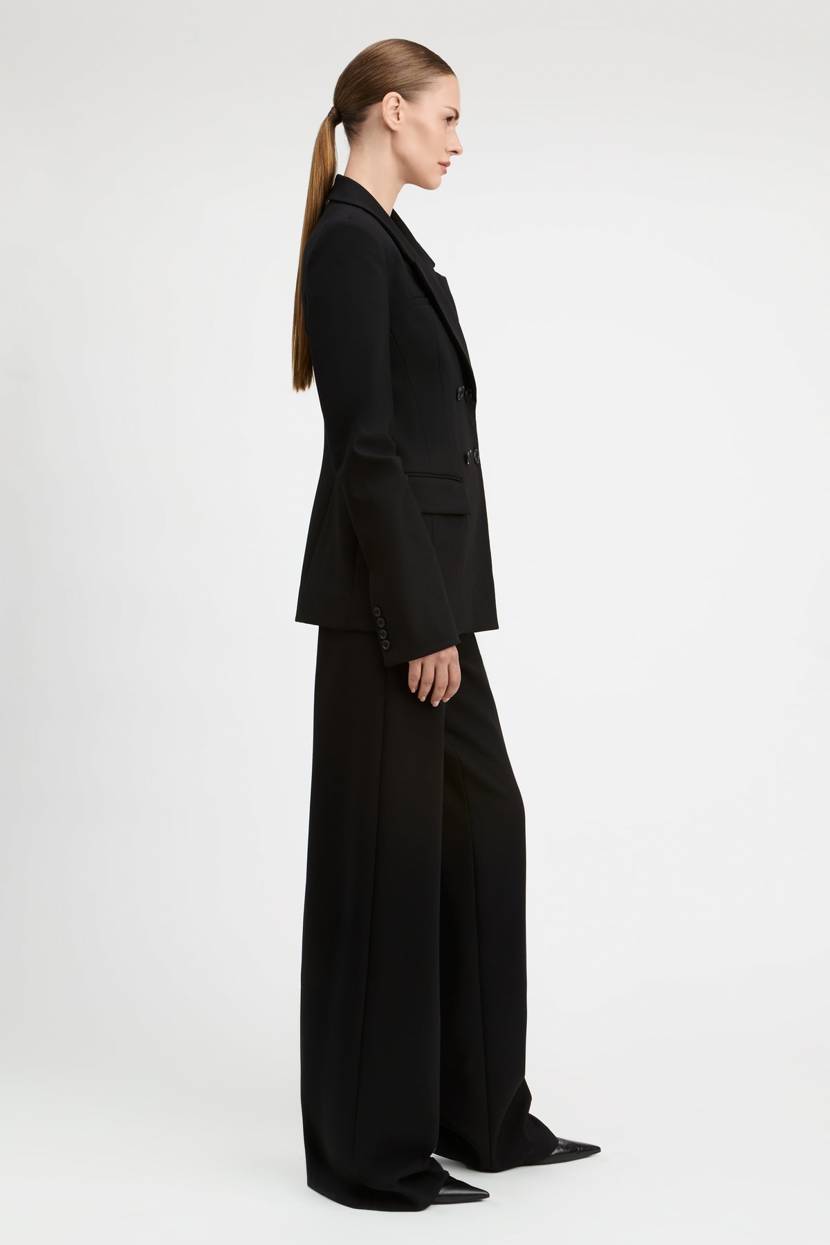 A model wearing a black double-breasted blazer and matching wide-leg trousers, photographed against a seamless light gray studio background.